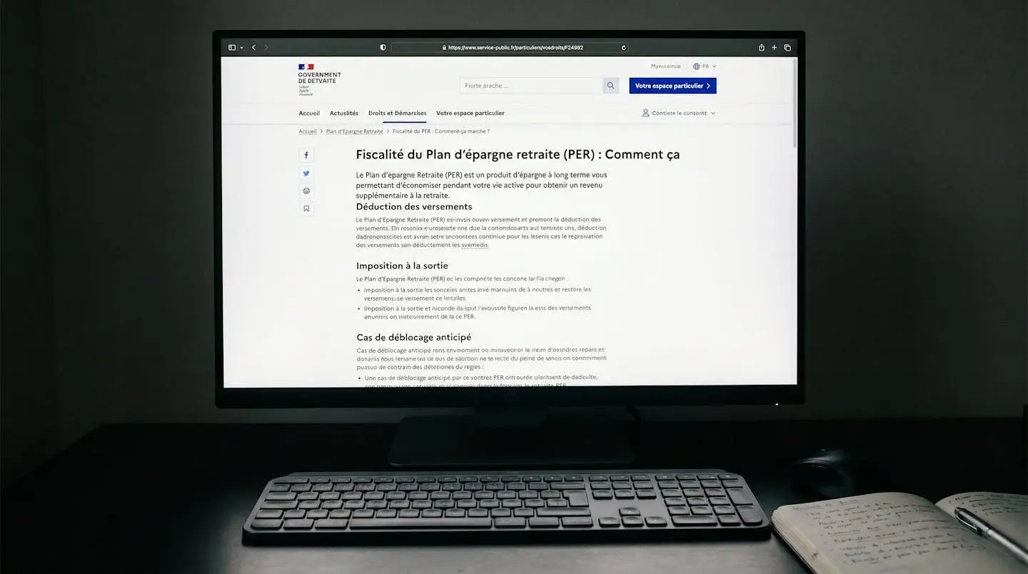 Écran d'ordinateur affichant page officielle service-public.fr ou impots.gouv.fr sur l'épargne retraite, avec clavier, souris et carnet de notes en premier plan, lumière d'écran douce